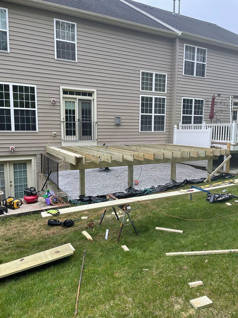 Deck construction in backyard: wooden frame, grey gravel, grass lawn, house with windows.