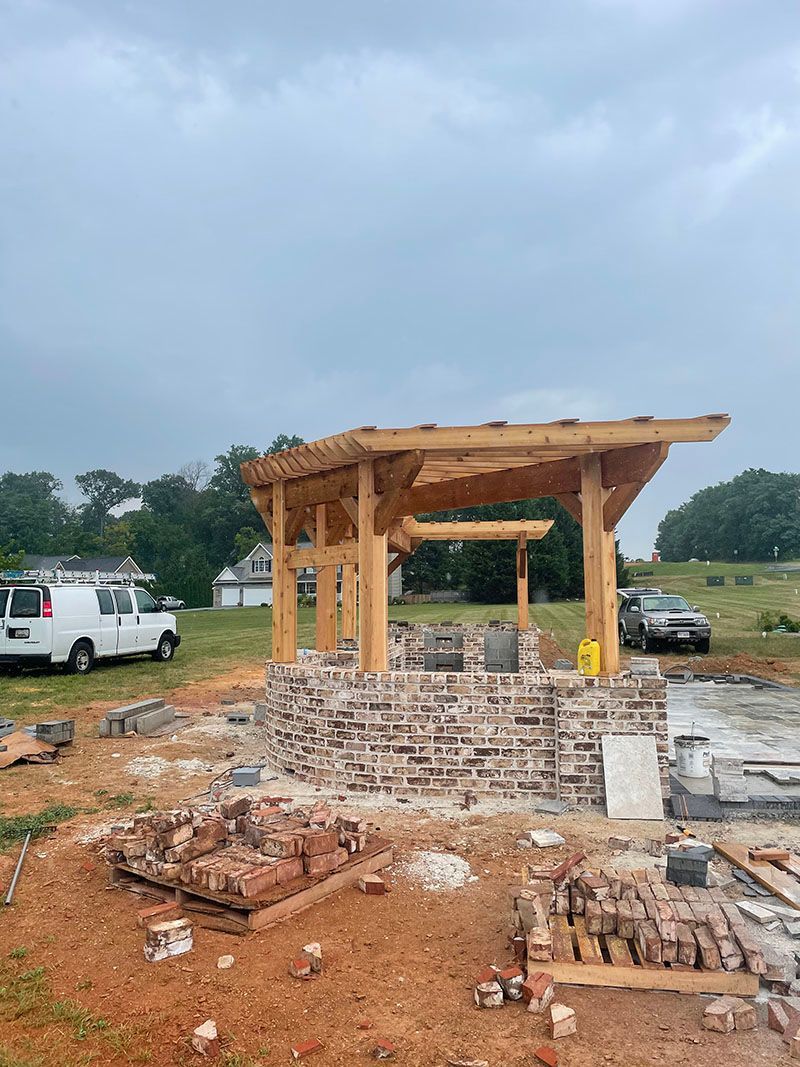 Wooden gazebo under construction, built atop a brick base, in a yard. White work vans nearby.