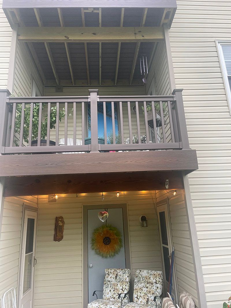 Two-story house exterior with a deck and a porch. Beige siding, brown trim, a blue door, and a sunflower wreath.