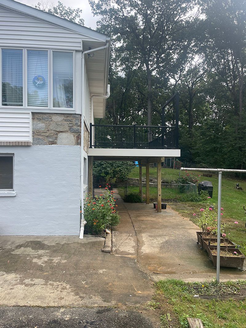 Side view of a two-story house with a small deck, concrete driveway, and overgrown yard.