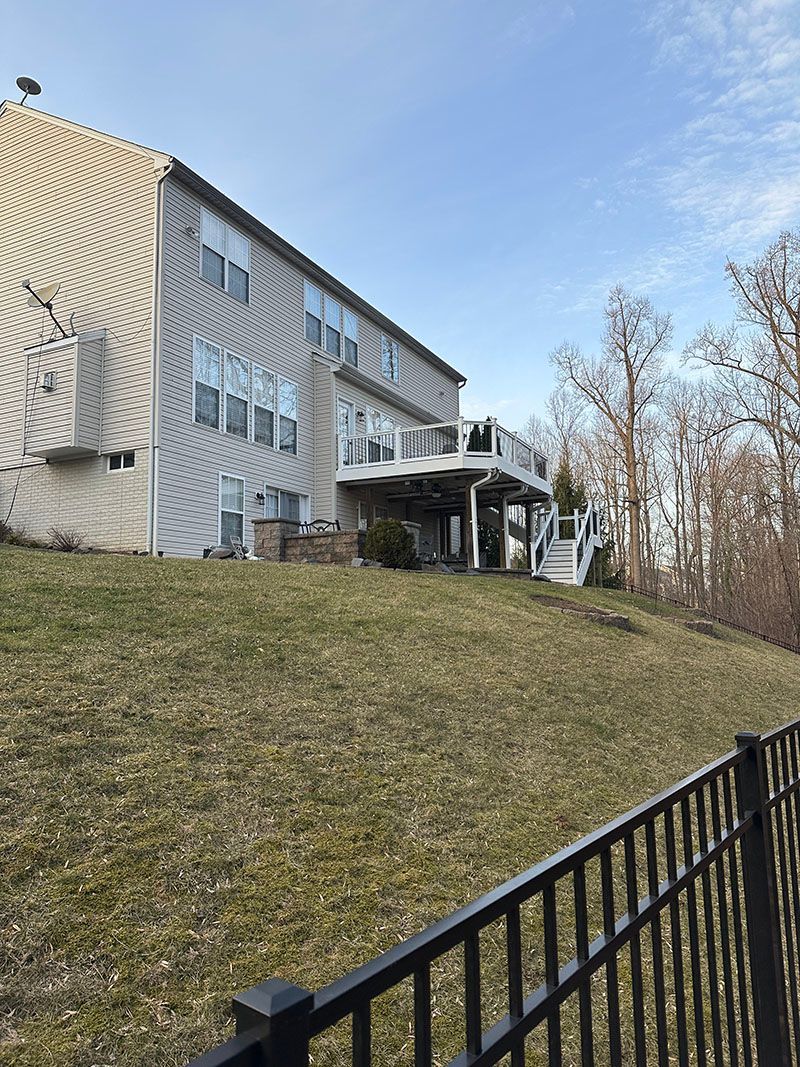 Two-story house with deck on a grassy hillside, black fence in foreground. Blue sky.