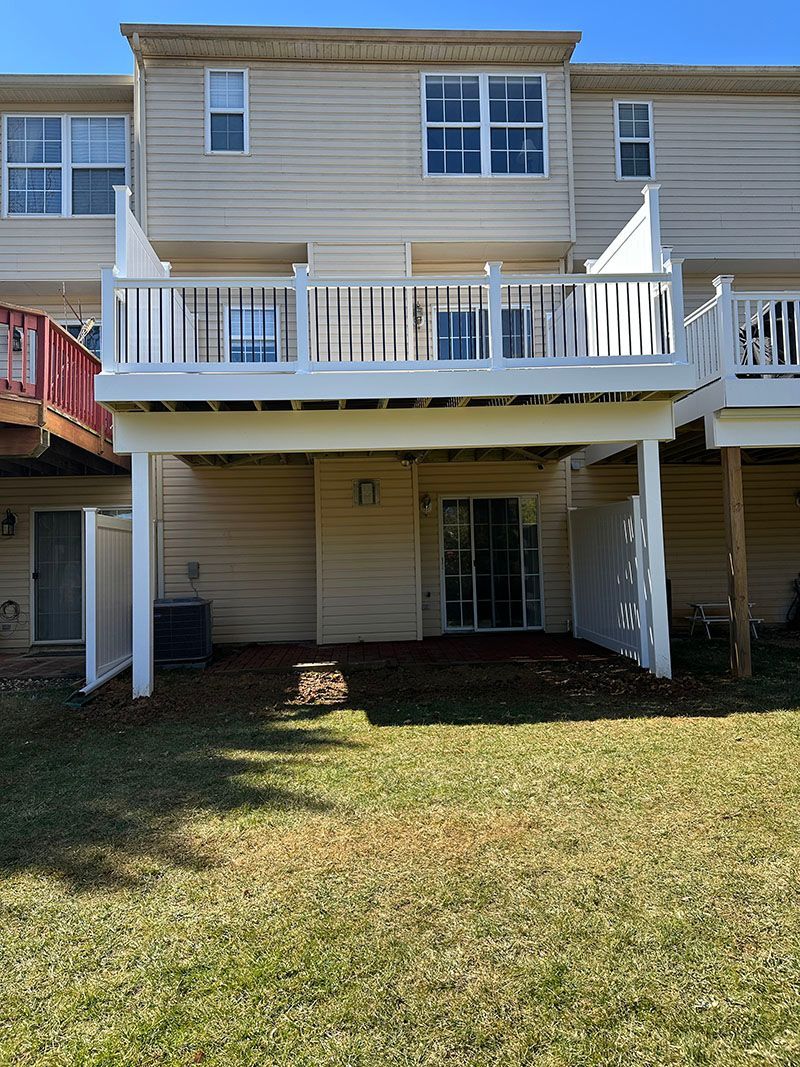 Back of a two-story beige townhome with a white deck. A yard with brown grass and a sliding glass door is below the deck.