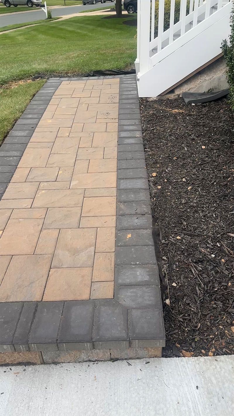 Brick walkway bordered by dark gray pavers, next to mulch and a white porch.