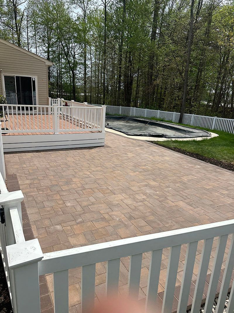 A large, paved patio with white fencing, deck, and a small pond in the background.