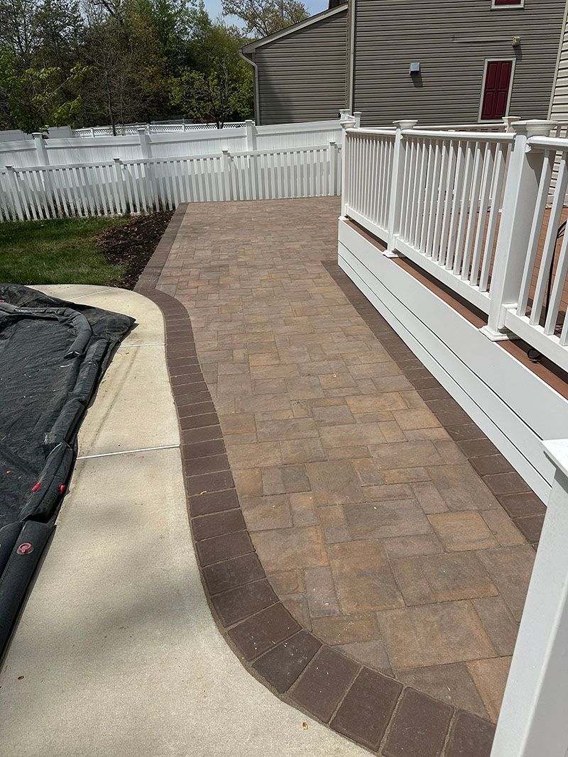 Brick pathway with a dark border, alongside a white fence and deck railing.