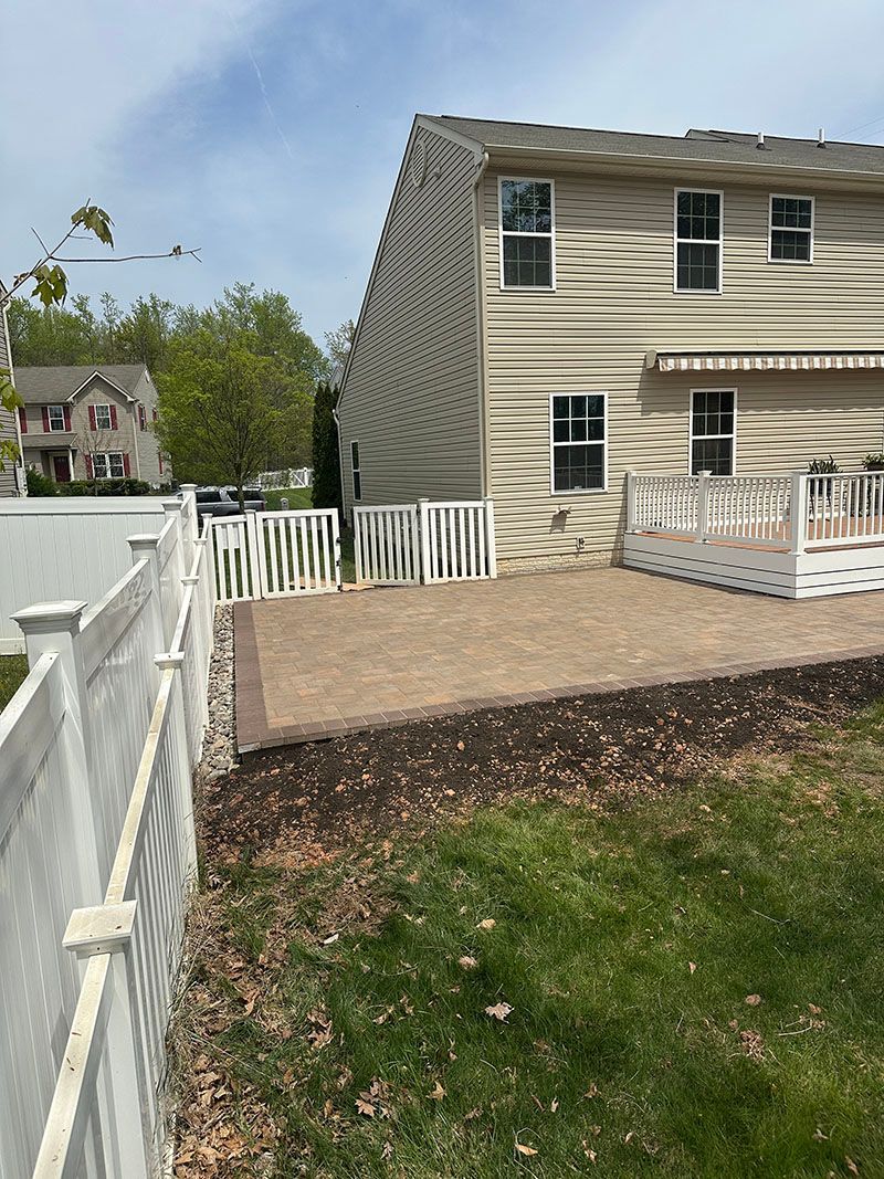 Backyard with a white fence, patio, and two-story house with beige siding. Green grass in the foreground.