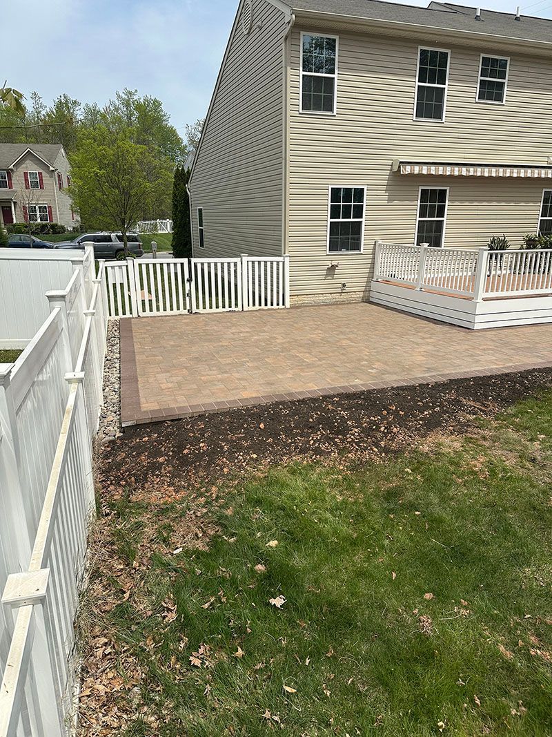 Backyard with a paved patio, deck, white fence, and a two-story beige house. Green grass and trees.