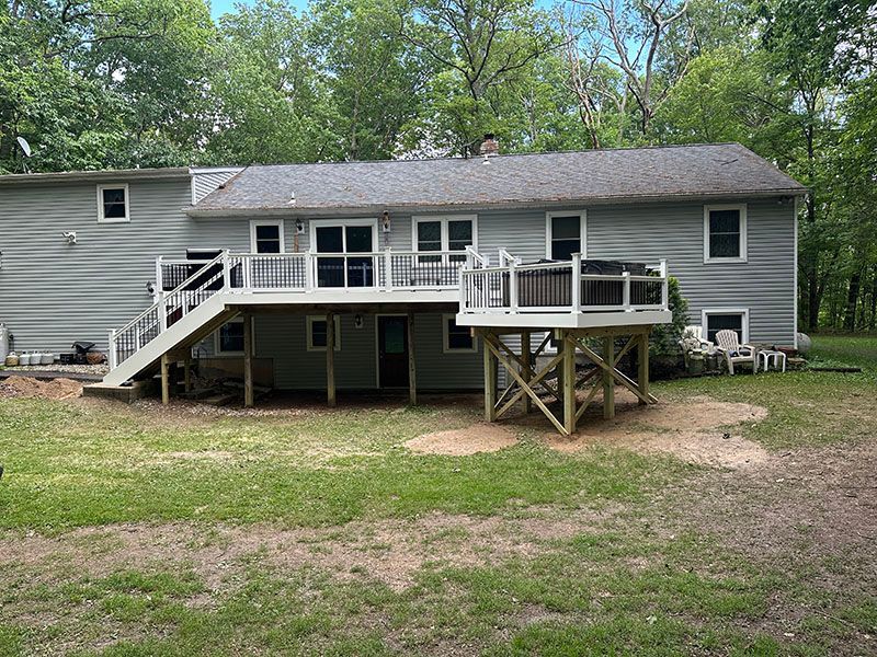 Back of a gray house with a white deck and a hot tub; trees in the background.