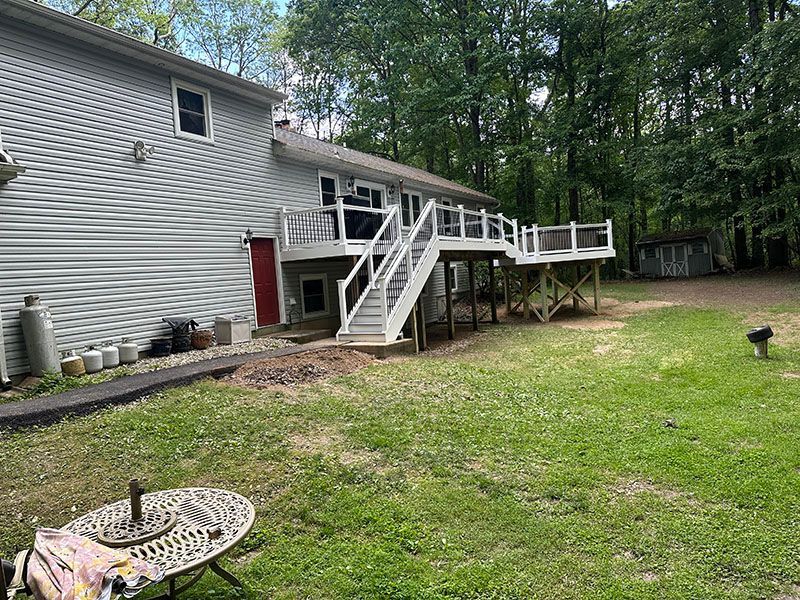 Two-story gray house with a raised deck and staircase. Green lawn, trees in the background.