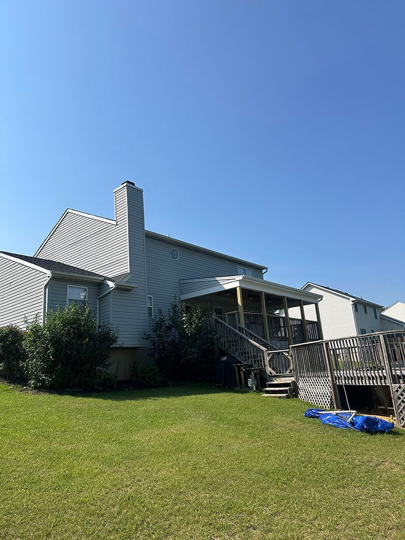 Backyard view of a blue-sided house with chimney, wooden deck, and covered porch against a blue sky.