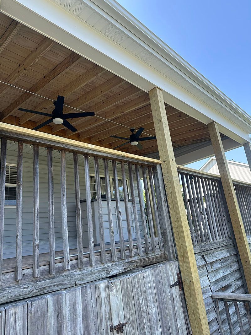 Wooden porch with ceiling fans, overlooking weathered deck and house. Sunny, blue sky.