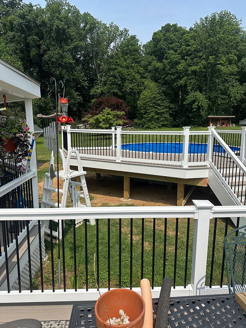 Backyard deck with a pool. White railing, green grass, trees, and blue pool water.
