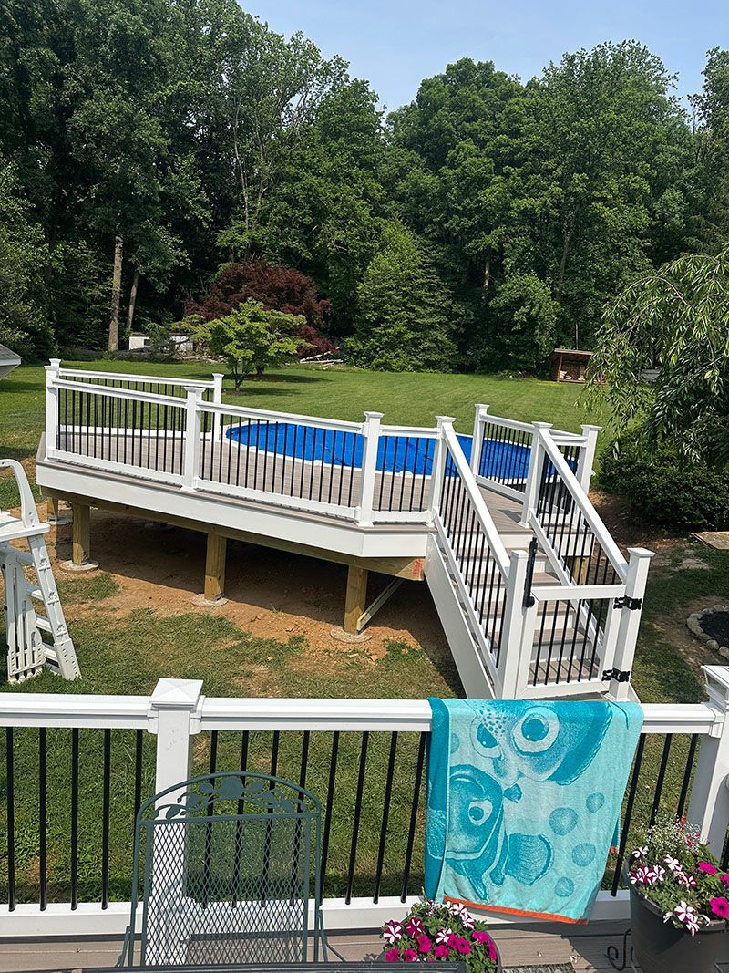 Above-ground pool with white deck and stairs, surrounded by trees, with a turquoise towel hanging on a lower deck.