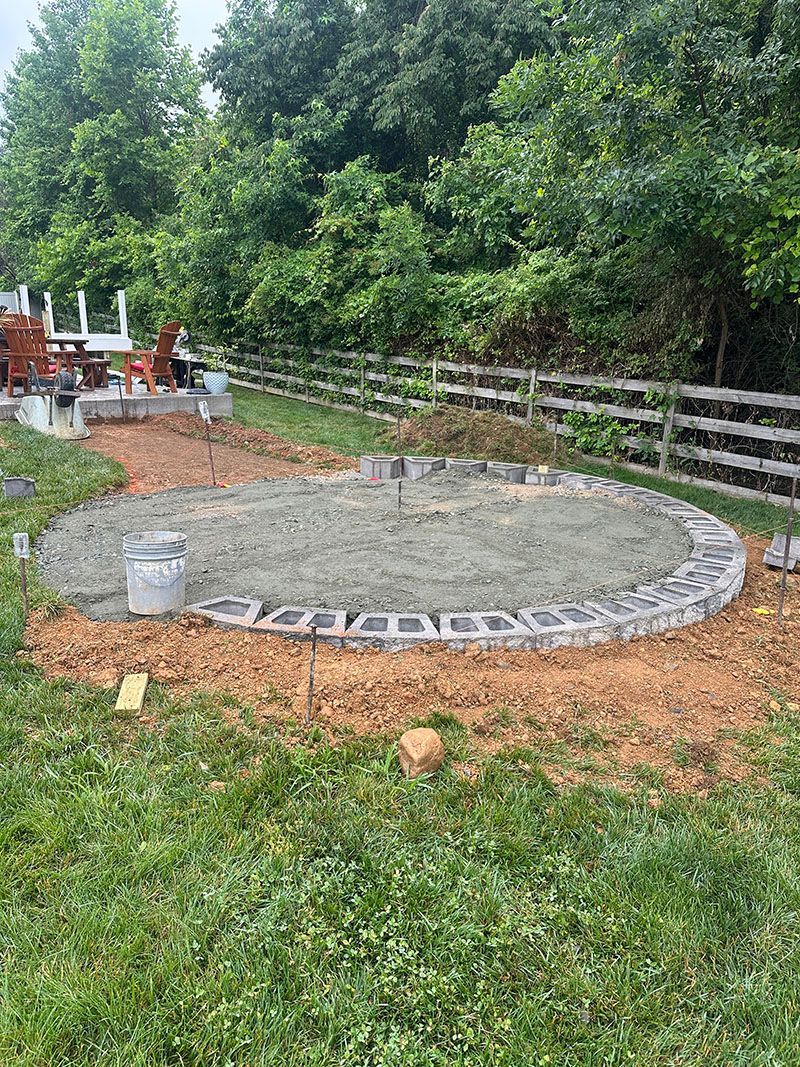 Circular patio under construction, bordered by gray blocks, filled with gravel, set in a grassy yard.