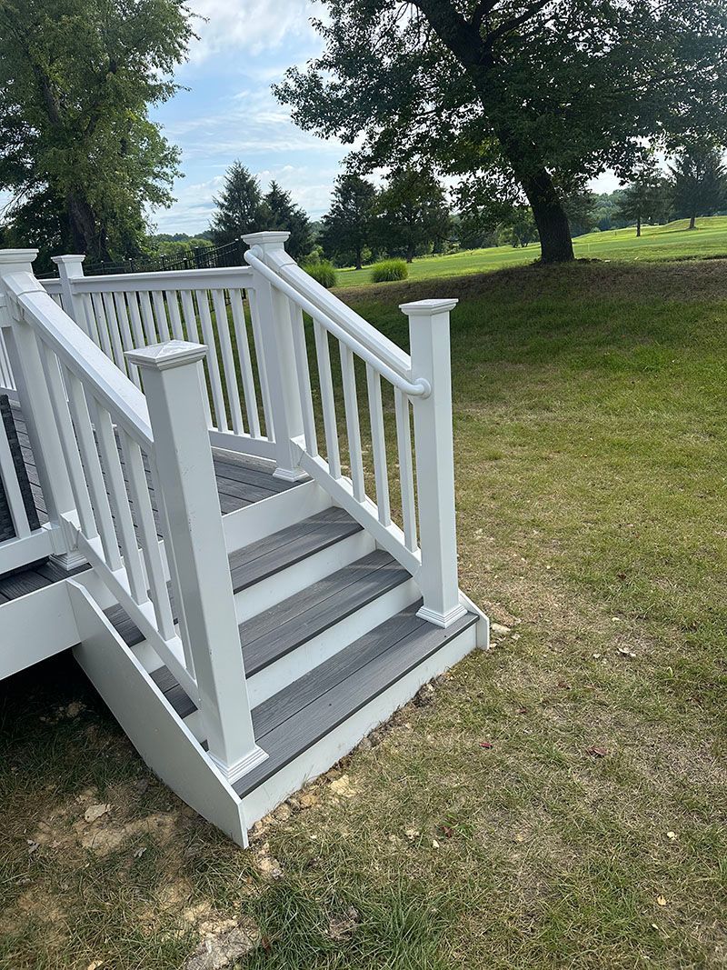 White deck stairs with gray treads, on a grassy hill, blue sky backdrop.
