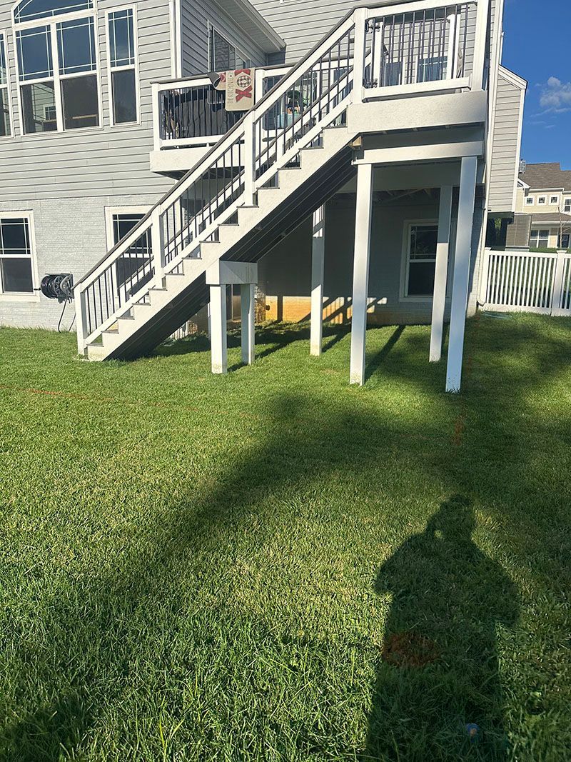 A deck with white stairs leads up to a light gray house. The grass in the yard is green and a shadow of a person is cast upon it.