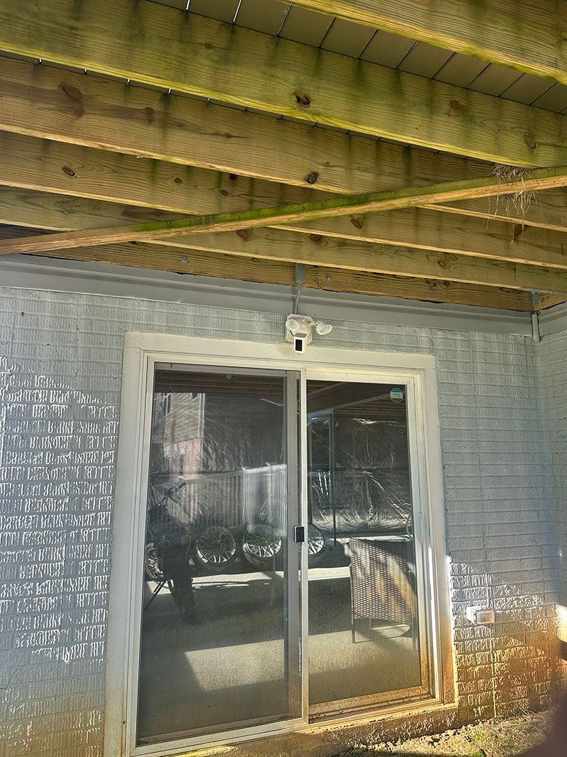 Sliding glass door under wooden deck, on brick wall. Security camera above door.