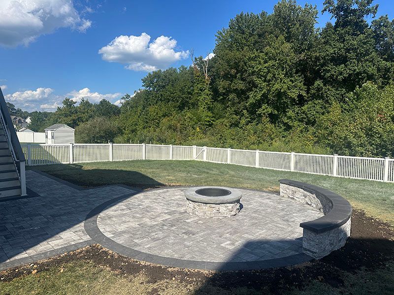 A backyard patio with a fire pit, surrounded by a curved bench, grass, and a white fence under a blue sky.
