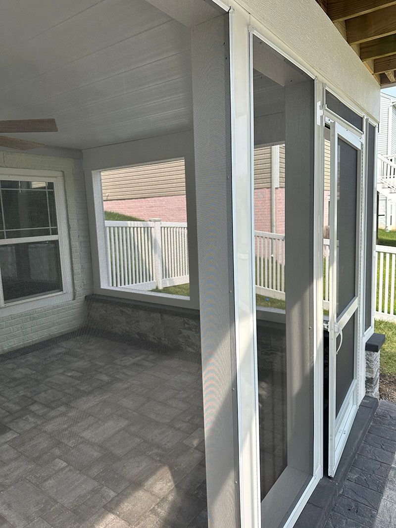 Screened-in porch with gray screens, white trim, and stone flooring. View of the yard and a white fence.