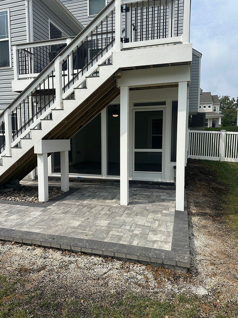 Brick patio under a raised deck, white supports, stairs, and a door.  Grey and brown bricks, grass and white fence.