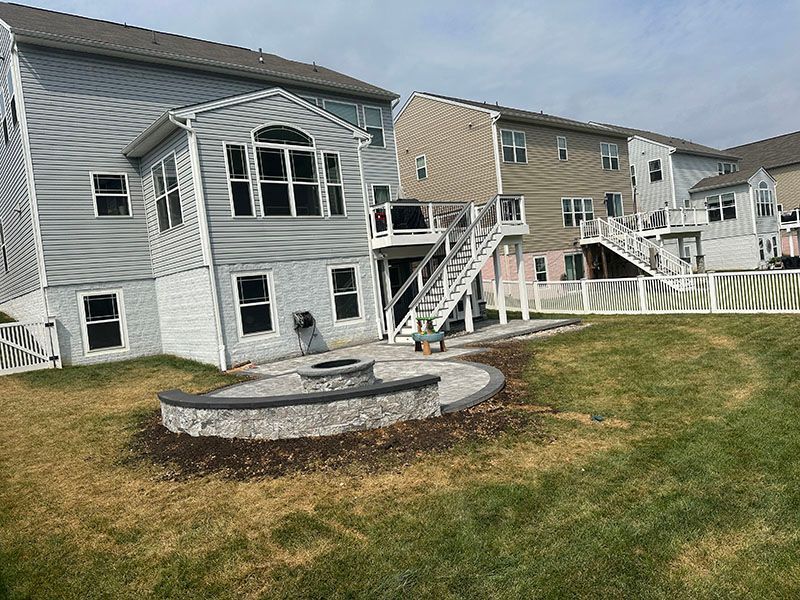 Backyard with house, deck, fire pit, and fence; green and brown grass, cloudy sky.