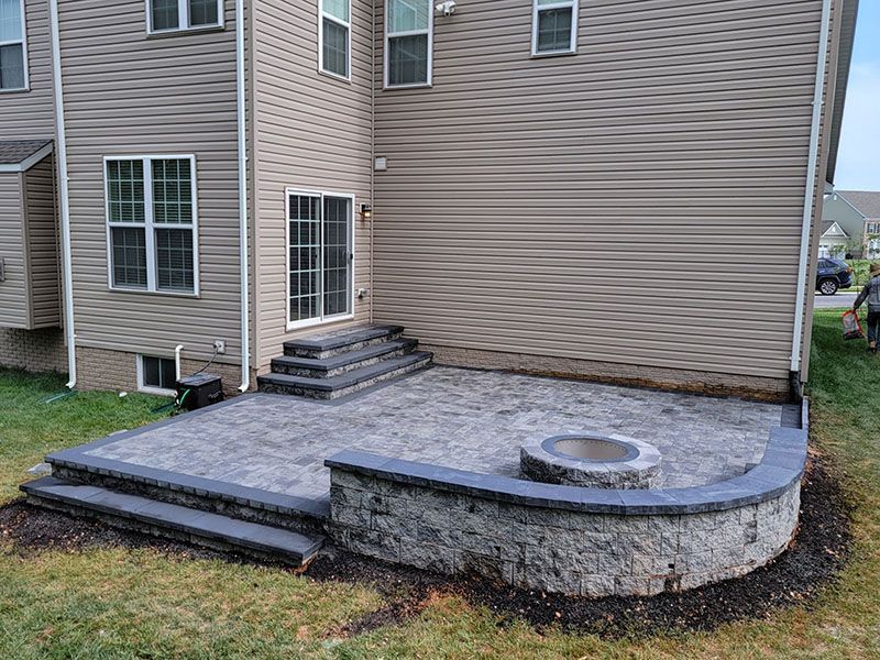 A gray stone patio with steps, a fire pit, and a retaining wall against a light-brown house.