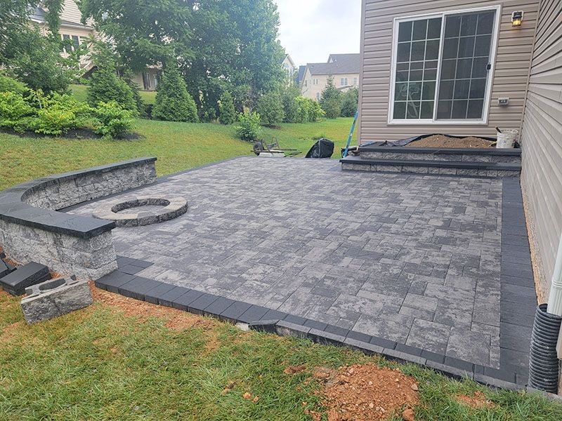 Patio with dark pavers, fire pit, steps, and bordering wall next to a house, surrounded by grass and trees.