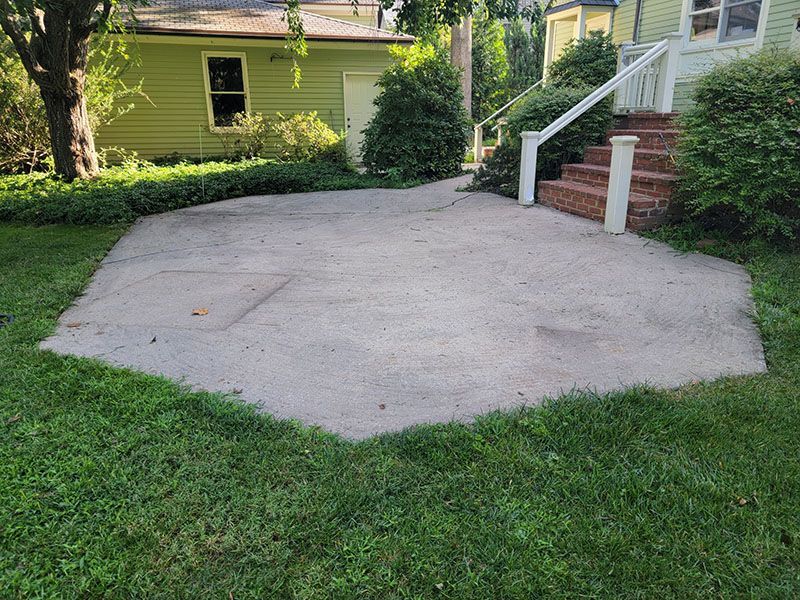 A gray concrete patio surrounded by green grass and bushes, with a green house in the background.
