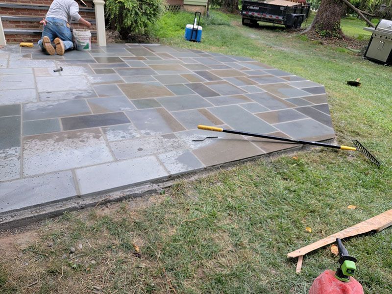 Person installing stone patio on a grassy area, kneeling and working.  Grey and blue stone, green grass, outdoors.