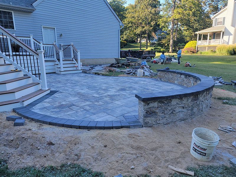 A brick patio with a curved stone wall being built next to a house and deck.