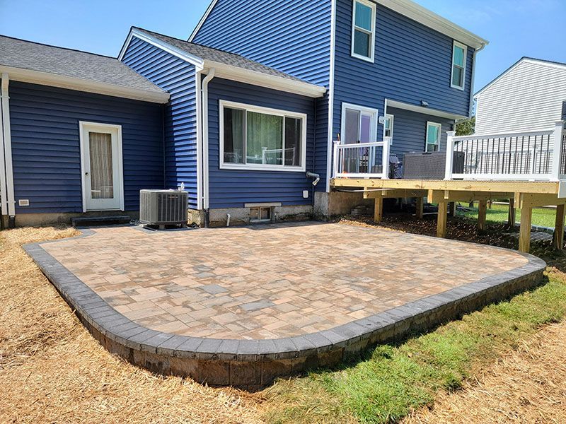 Paver patio bordered by gray blocks next to a blue house with a wooden deck and lawn.