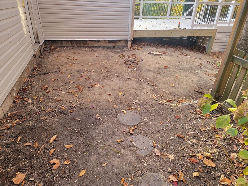 Dirt area with fallen leaves, next to a white-sided building and a wooden deck with railing.