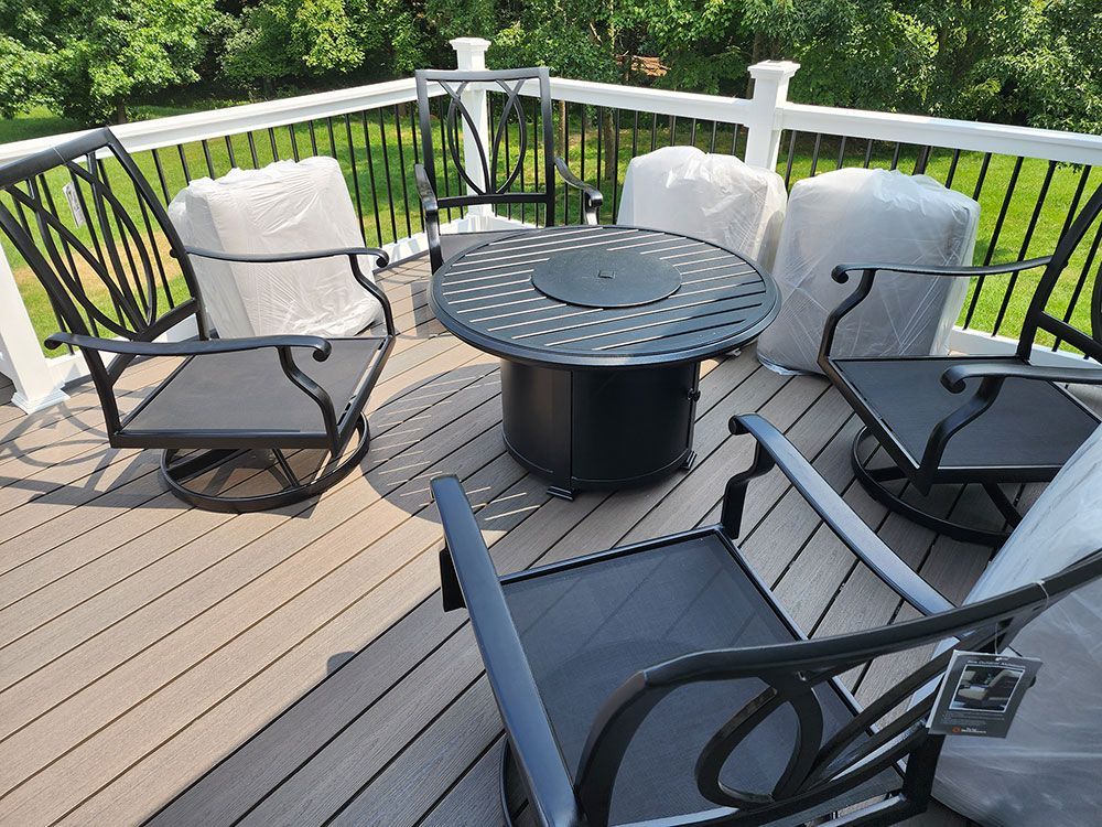 Patio set on deck: black chairs, round table, fire pit. Chairs are covered. Green trees in background.