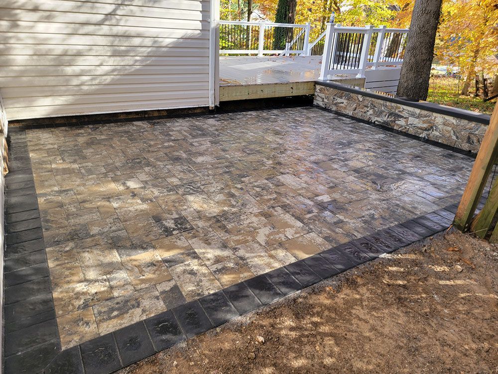 A patio with tan and grey pavers, bordered by dark pavers, next to a white house and a stone wall.