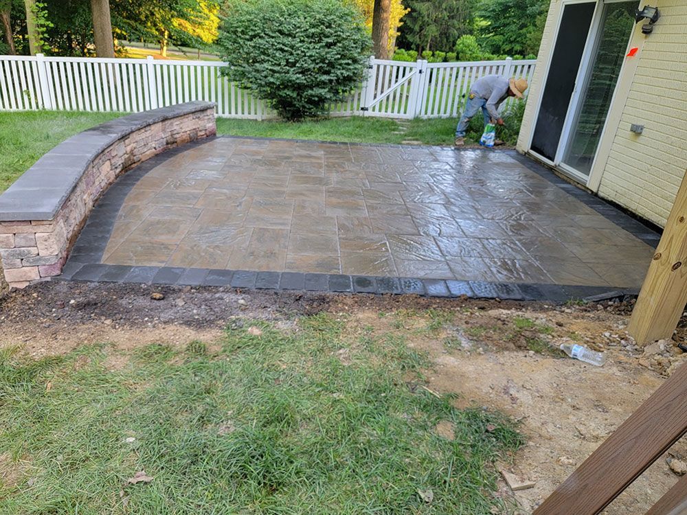 Man cleaning a newly paved patio with a low brick wall and white fence in the background.