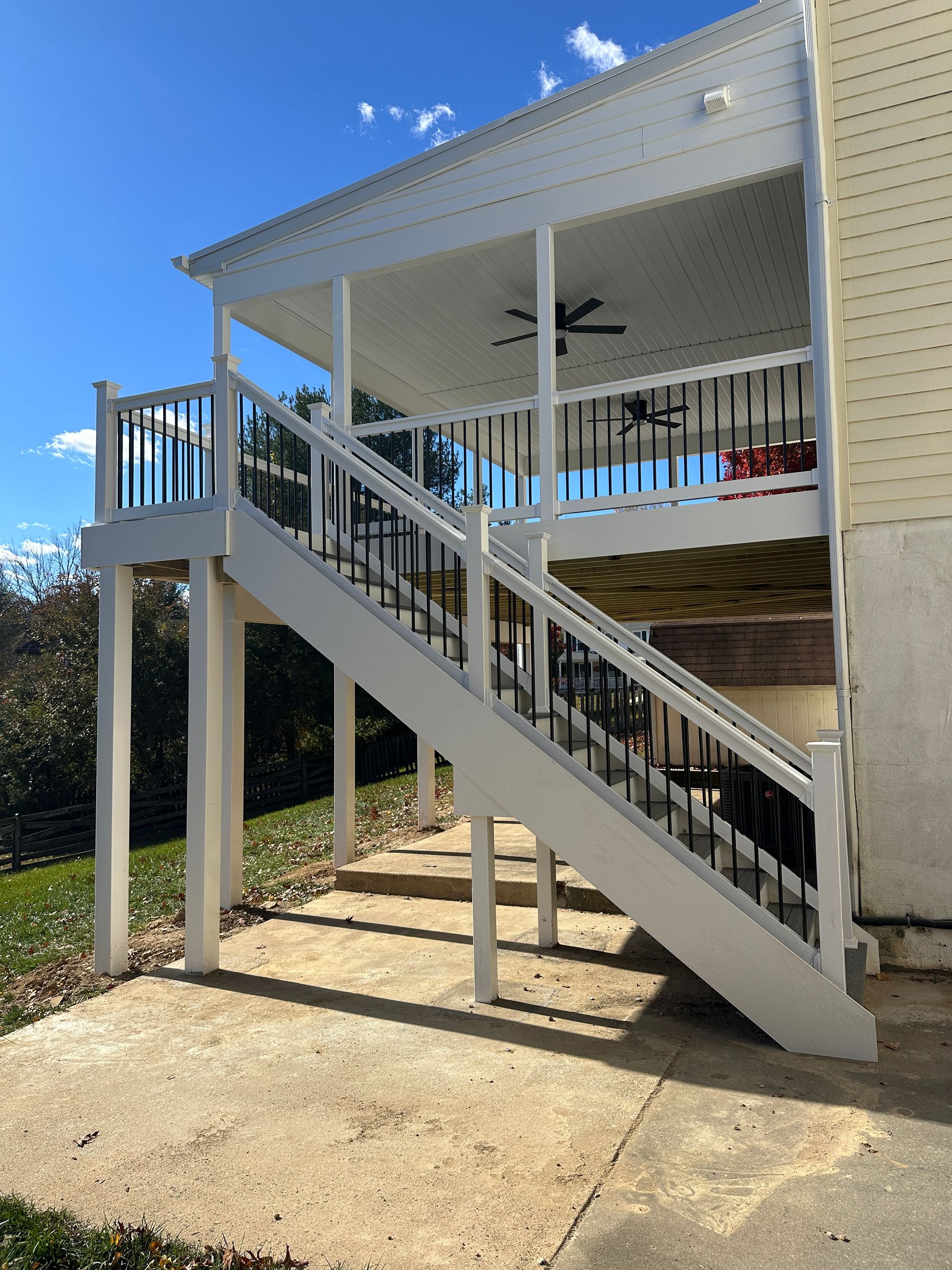 White elevated deck with stairs and black railings against a yellow house, on a concrete patio, sunny.