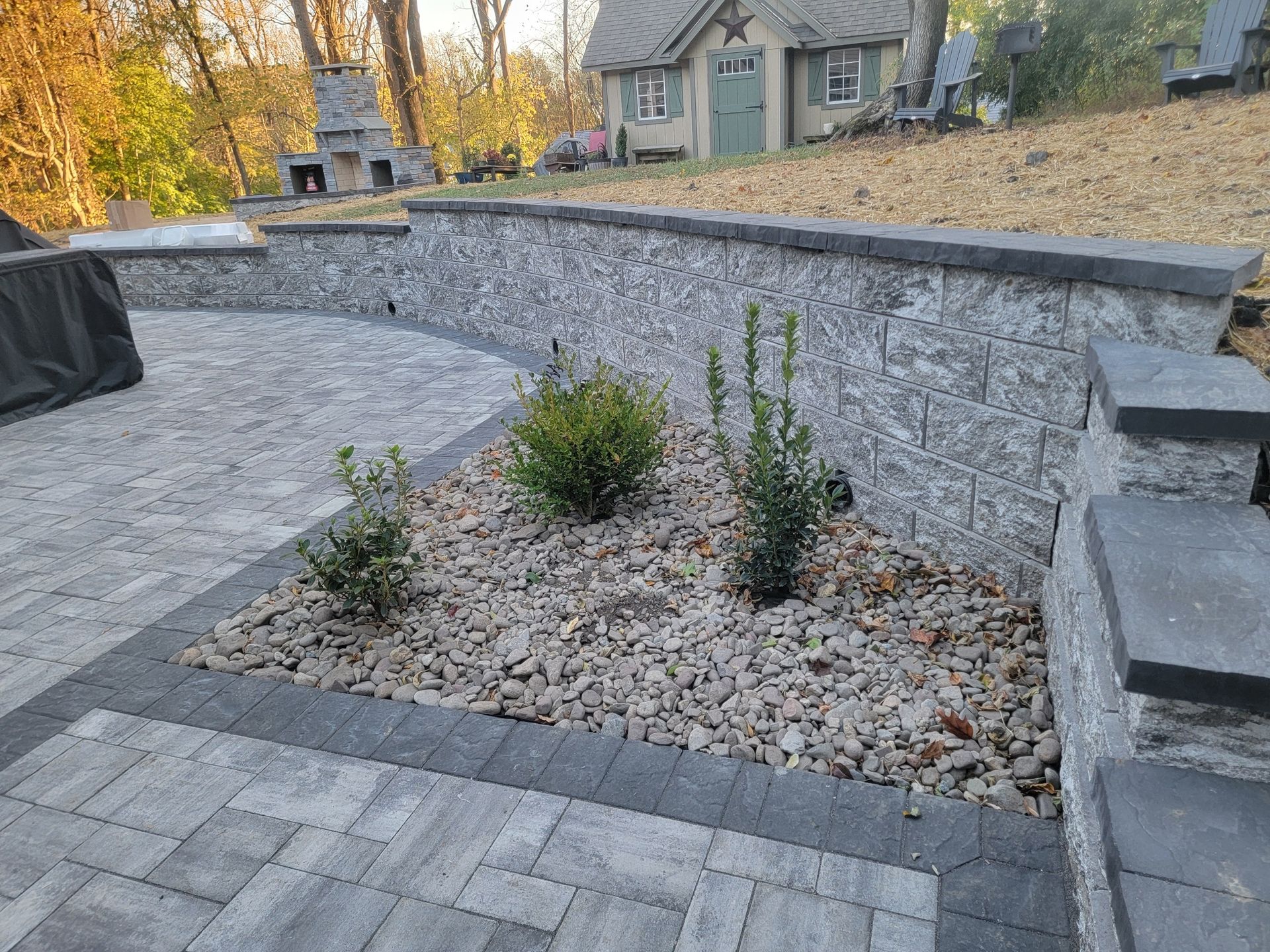 Patio with a stone wall, small bushes, and gravel. A house sits in the background.