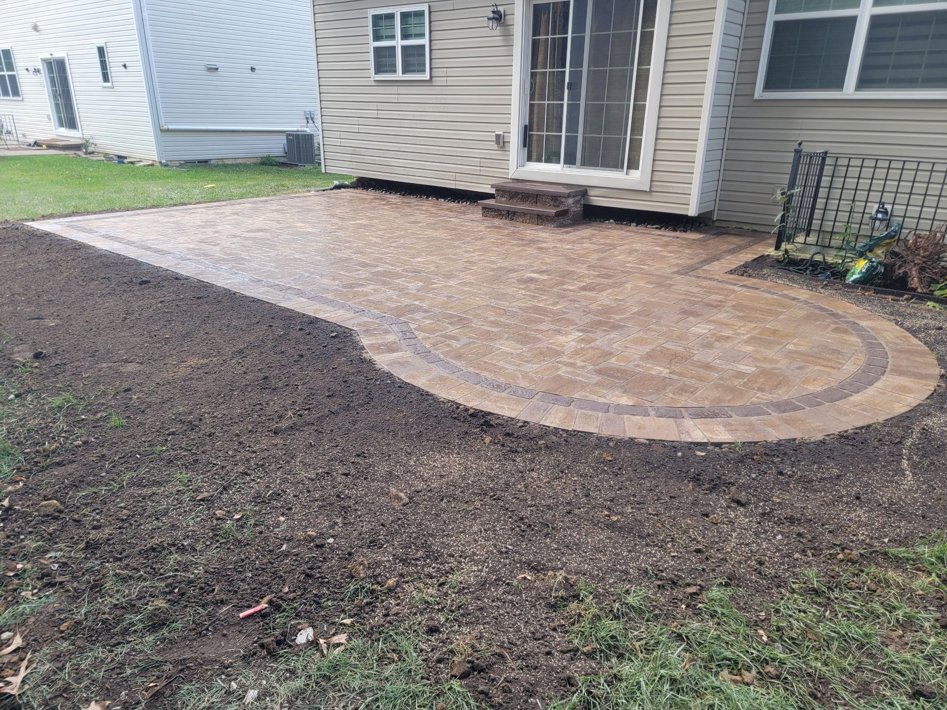 Stone patio with curved border, adjacent to a house, surrounded by dirt and grass.