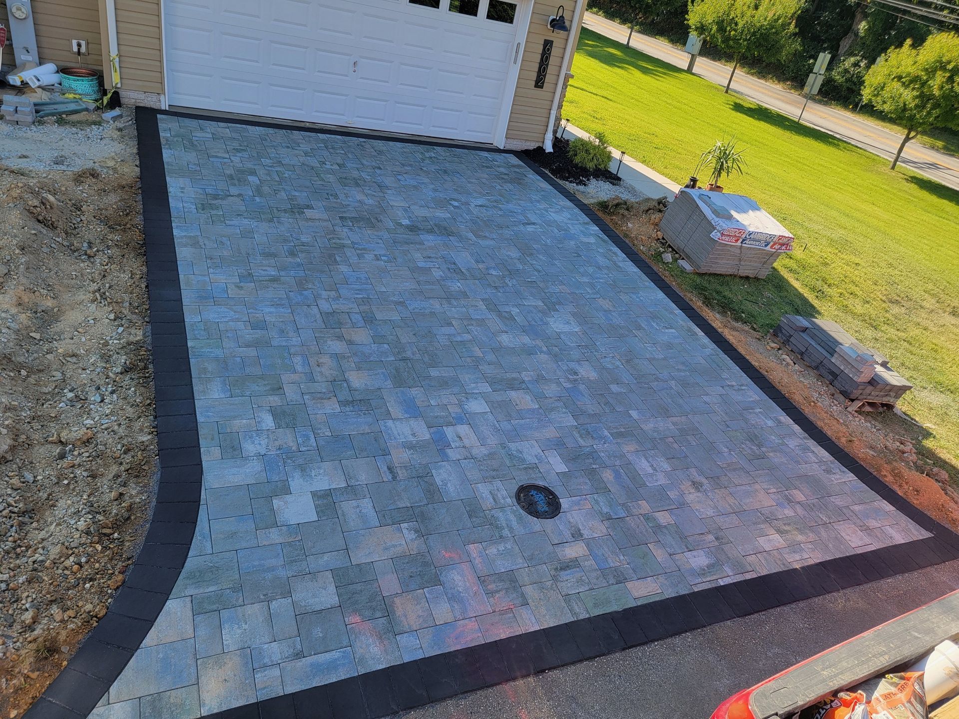 Driveway paved with gray and black bricks, in front of a garage, next to grass.