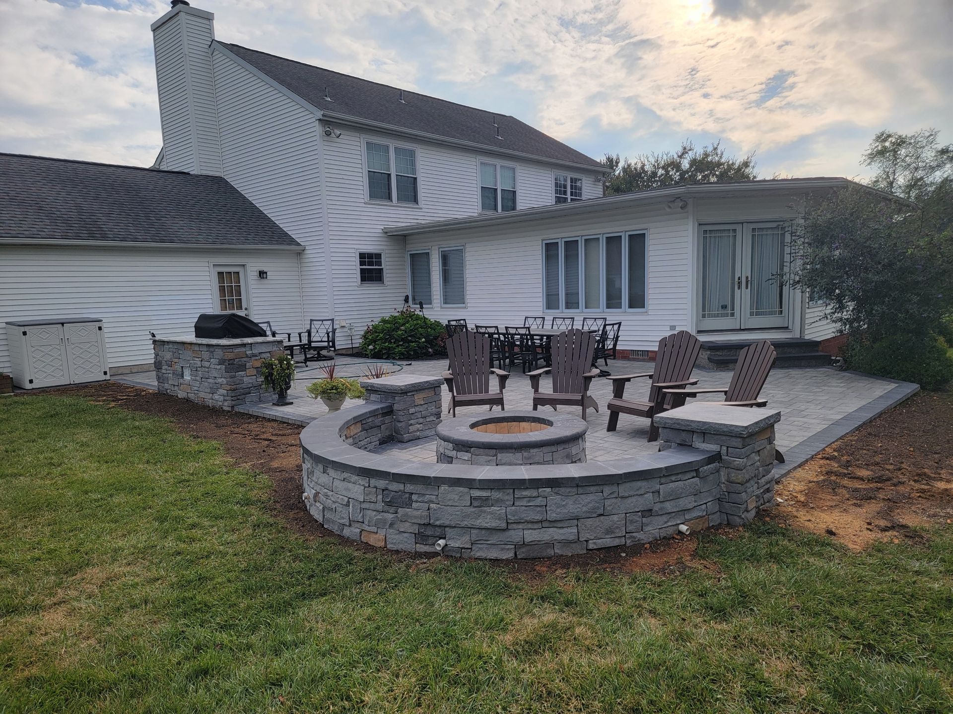 Backyard patio with fire pit, seating, and house in the background. Gray stone and lawn present.