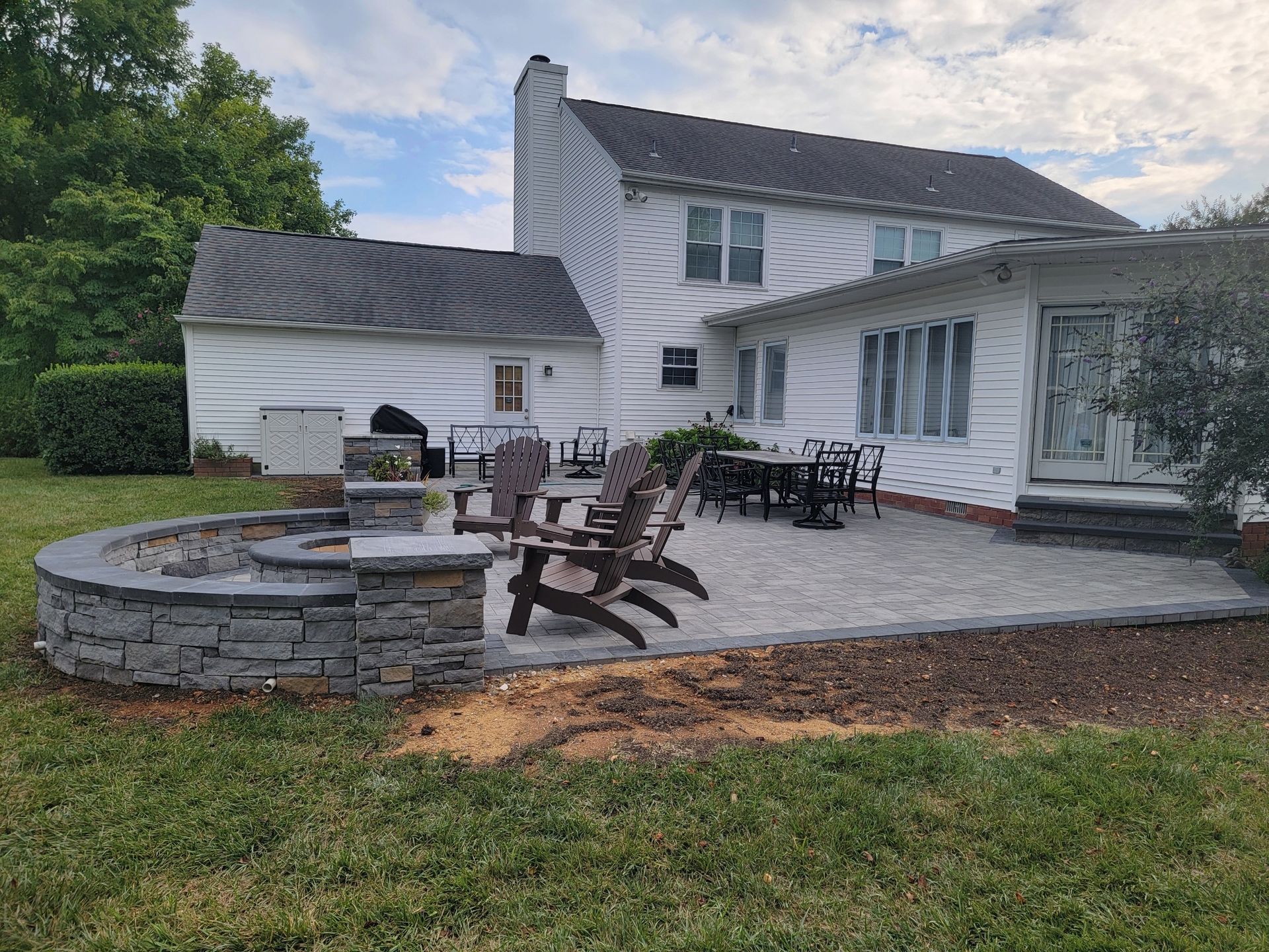 Backyard patio with fire pit, seating, and house in the background. Gray pavers and stone with dark wood chairs.
