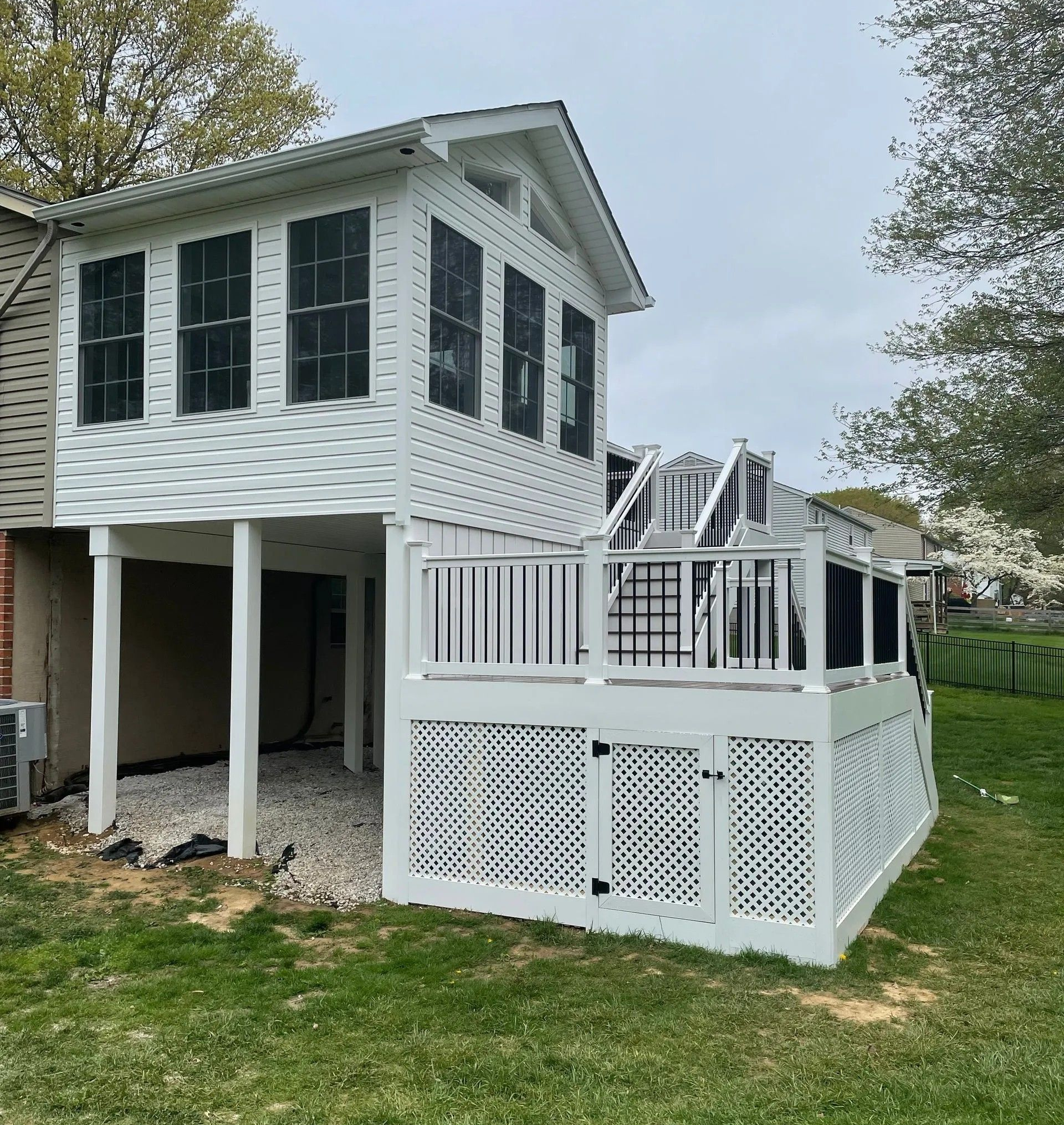 White two-story deck addition with lattice, stairs, and black shutters; seen from a grassy yard.