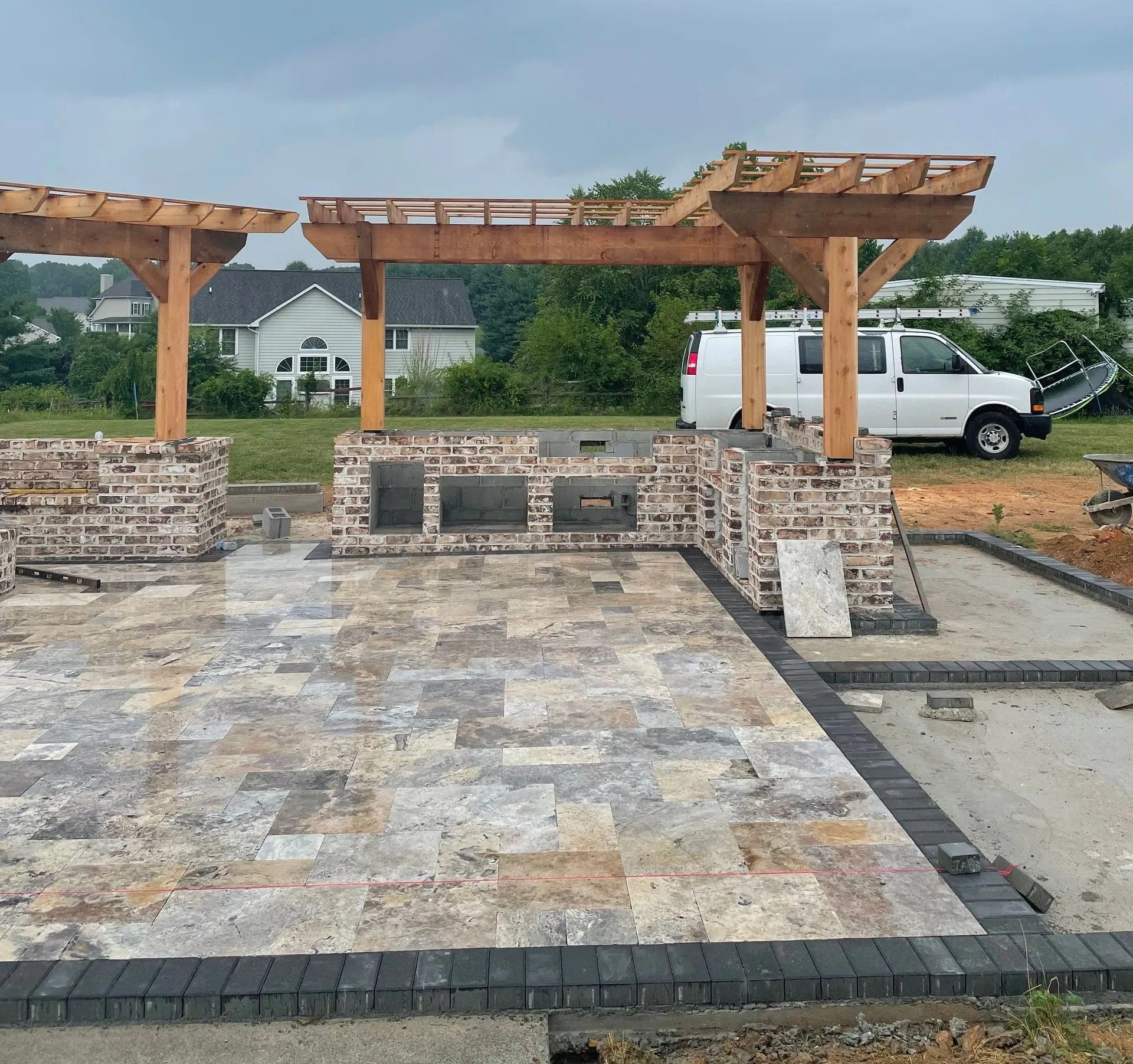 Construction site with pergola framework over stone grill, paved patio. Van parked nearby. Cloudy sky.
