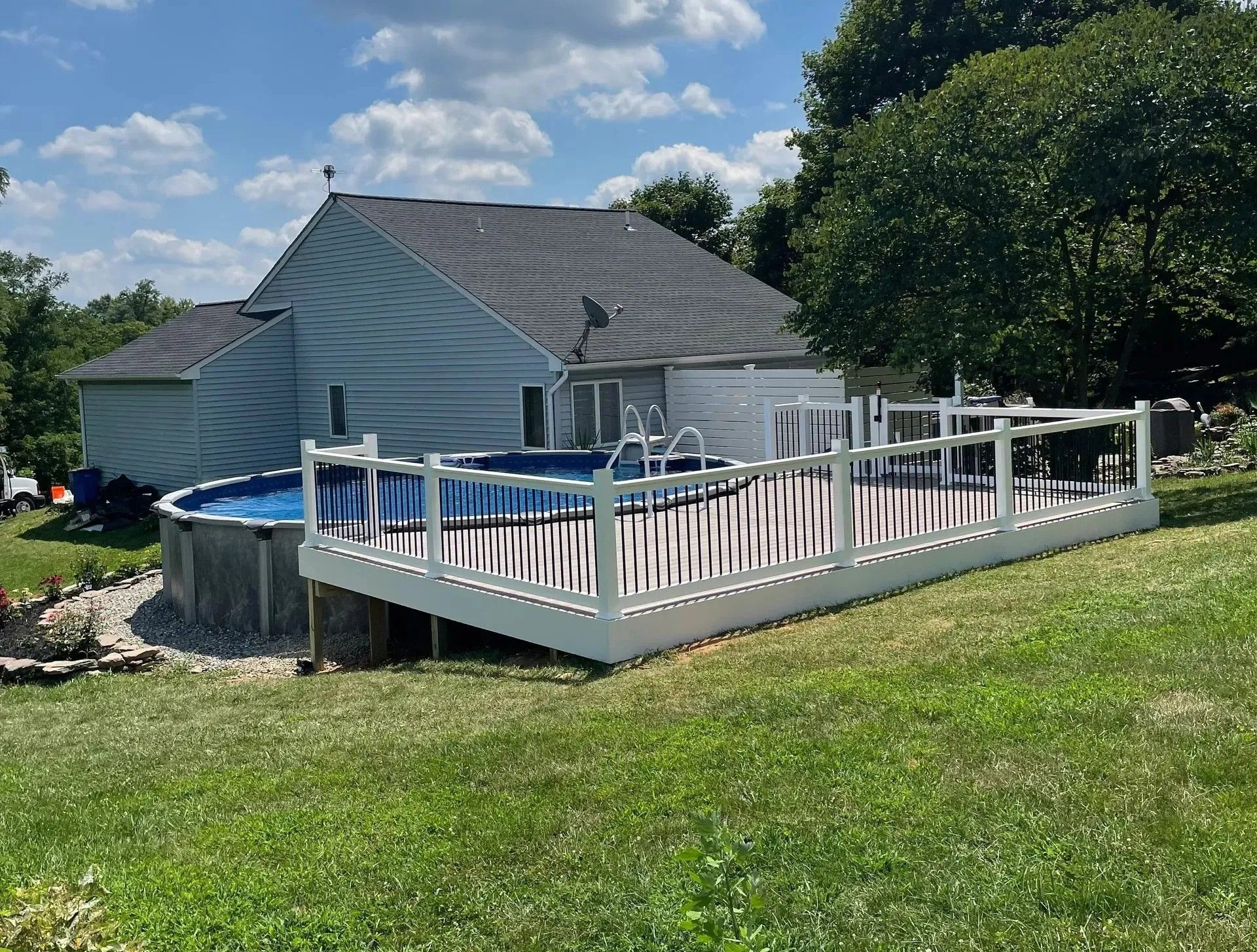 White deck surrounds above-ground pool next to a house on a sunny day with blue sky and clouds.