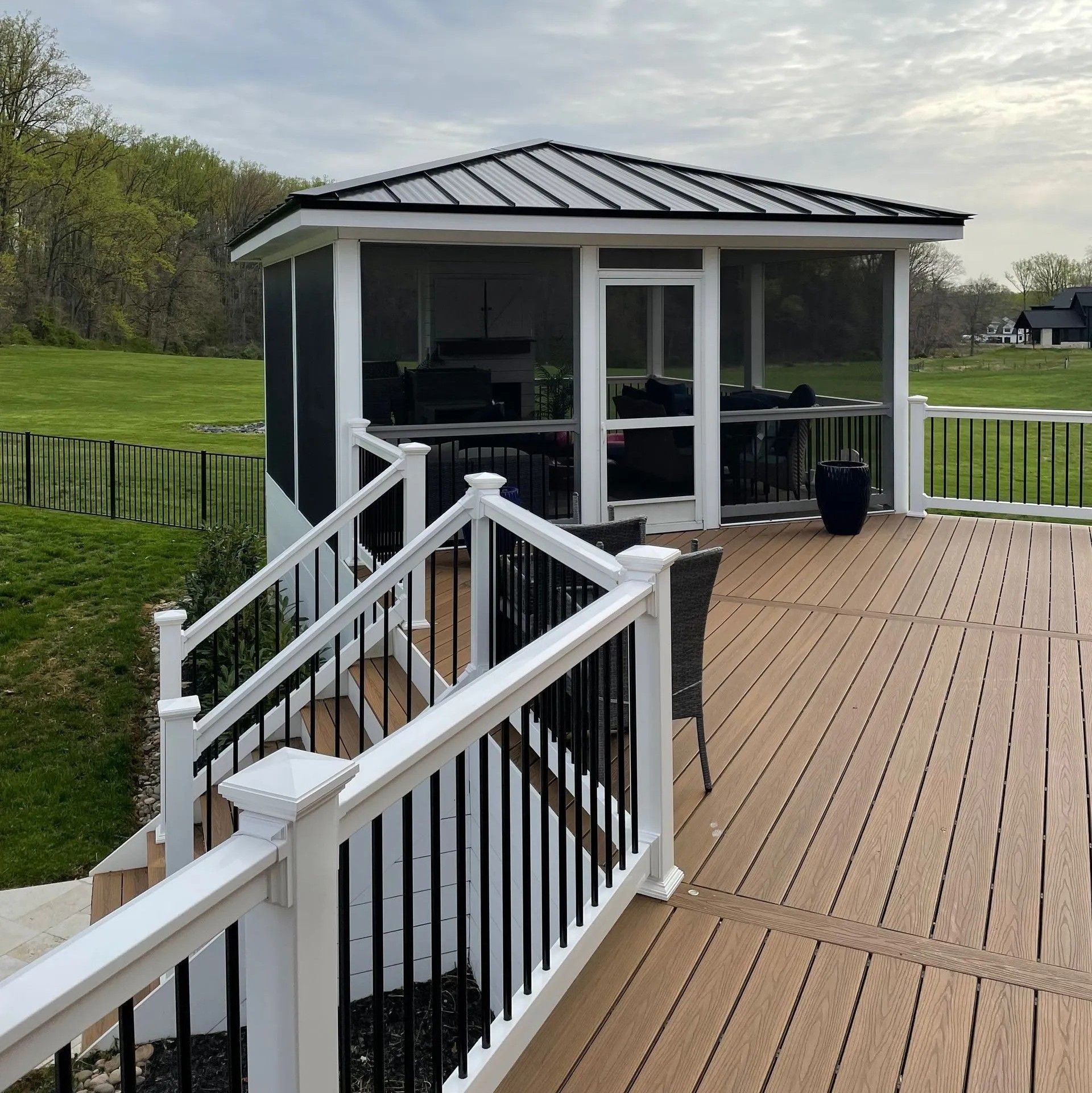 A screened gazebo on a wooden deck with a view of a green field and cloudy sky.