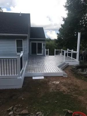 A large gray wooden deck attached to a light blue house, surrounded by a grassy yard.