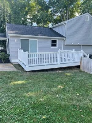 A white deck attached to a gray house, surrounded by green grass and trees.