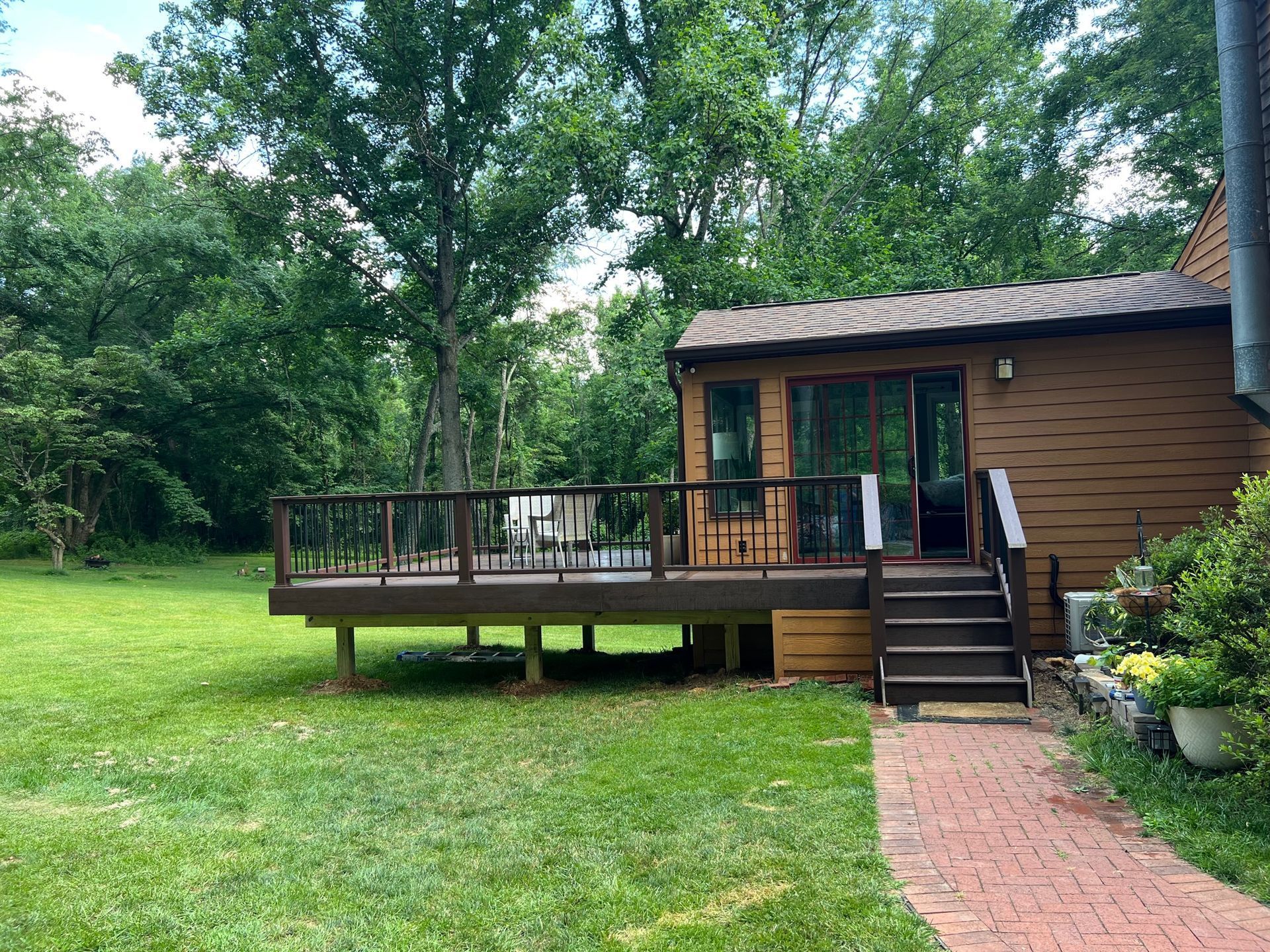 Brown house with a deck, steps, and brick path in a grassy yard surrounded by trees.