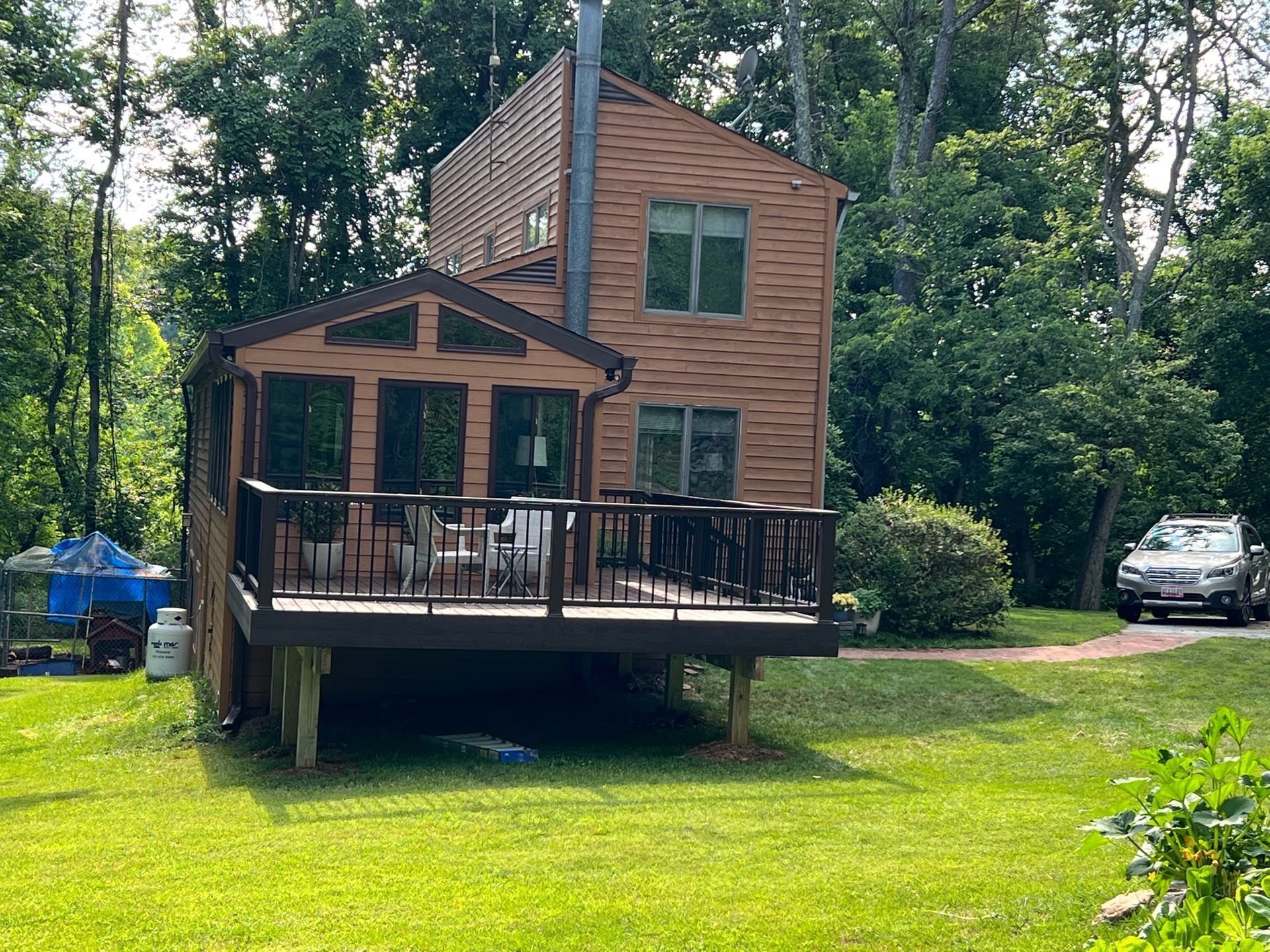 Brown house with deck, porch, and car in the yard, surrounded by trees on a sunny day.
