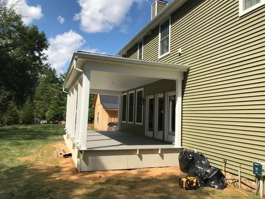 A light-colored covered porch attached to a green-sided house, on a sunny day.
